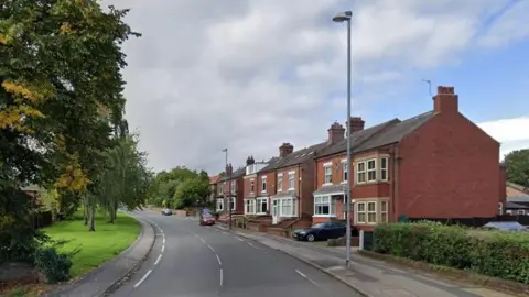 A street view image shows a row of red brick houses on the right and trees to the left of the road. 