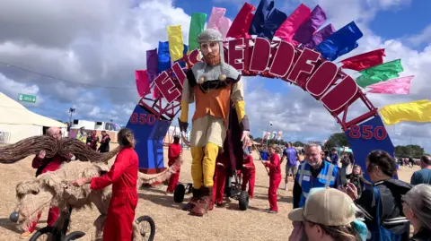 Scene at Eisteddfod with arch spelling out Eisteddfod and 850 either side to mark the festival's anniversary. A very large puppet is visible under the arch with people in red jumpsuits behind moving it along with the help of wheels as people look on. A smaller dog puppet on wheels is visible in front.