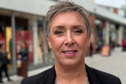 Sharon Edwards/BBC Lisa Fitzgerald stands in front of modern shop fronts at the Pescod Square shopping precinct. She has short hair and wears a smart black jacket and a silver necklace.