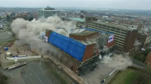 Brick-built bus station, partly covered in blue sheeting, with a large cloud of dust on the left side following the detonation of demolition explosives. There is a multi-storey building to the right and a car park to the left.