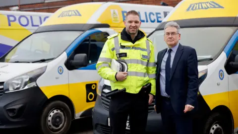 A uniformed male police officer in a hi-viz yellow jacket, holding his cap under one arm, stands next to the commissioner, with greying hair and wearing a dark blue suit and tie. They are in front of a fleet of speed enforcement vans with police livery and speed awareness messaging.