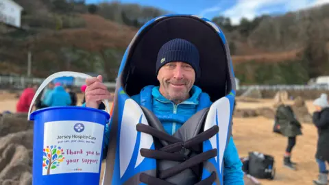 A man wearing a blue beanie and a blue shoe costume. He is stood on a beach and is holding a fundraiser bucker which says Jersey Hospice Care. 