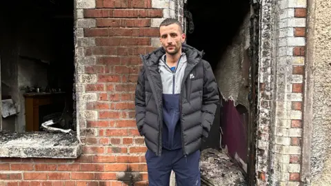 A man with brown hair and a black puffer coat stands outside his home 