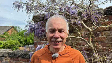 Caradoc with grey hair and a moustache wearing an orange hoodie and smiling at the camera. Behind him is a wall with purple flowers growing on it.
