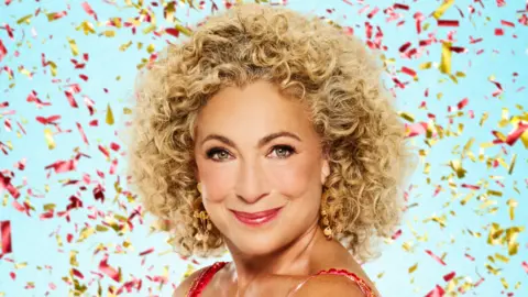 Alex Kingston has curly blonde hair and is standing in front of a blue wall whilst confetti falls behind her.