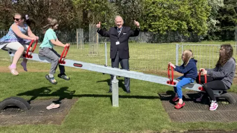 Four children on sea saw, with a man in the middle, with his arms up, wearing a black suit. The park has lots of green grass and trees. 