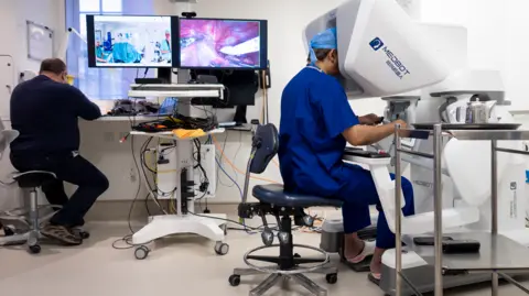 Doctor sits looking into a white box whilst another man sits in front of two screens showing the surgery from two different angles.
