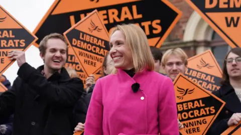 PA Media Woman wearing a pink coat smiling in front of orange "Liberal Democrat" campaign boards and happy-looking supporters