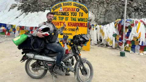 We Hate The Cold Harry Jackson poses on a moped, with his left thumb up. There are rocks behind him and a large handpainted sign featuring the name and altitude of the mountain pass, along with some colourful flags. He is pointing to the sign, wearing boots, trousers, a jacket, holding a helmet and wearing a jacket. 