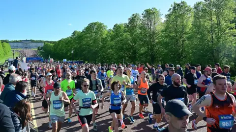 Pacemaker Press Numerous runners take off from the marathon start line wearing various coloured sportswear. There are spectators along the side of the road and Stormont in the background.