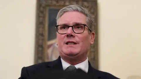 Keir Starmer, wearing dark-rimmed glasses and a dark suit, looks off camera, mid-speech 