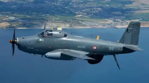 A grey military plane with the word marine on its side flies over the sea, with a beach and town in the background. It is a bright day.