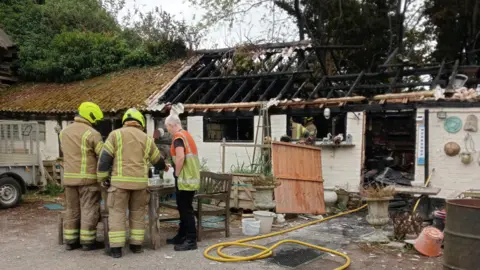 Contributed Firefighters wearing uniform and helmets stand outside a building that has been destroyed in a fire. They are gathered together, talking, with their backs to the camera. The building's roof, windows and inside has been destroyed.