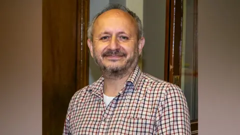 A man with grey hair and a grey beard, wearing a red and blue check shirt. 