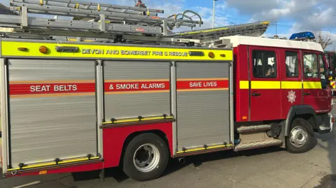 A Devon and Somerset Fire Service engine parked on a road. It has several ladders visible on its roof. It has a message saying "Seat belts and smoke alarms save lives" on its side.