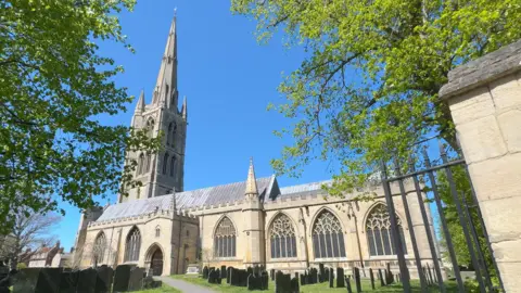 A large historic stone church with a tall spire stands behind a graveyard, viewed through an open gate. The church is framed by leafy trees under a clear blue sky, with gravestones and a paved path visible in the foreground leading towards the building.