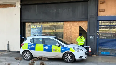 A police car parked outside a boarded shop. There is a police officer standing behind the car.