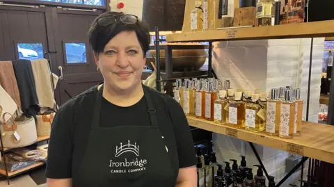 Cherryl Montrose-Matheson standing in her shop wearing a black top and black apron with Ironbridge Candle Company. She is standing in front of a shelf of products and is looking into the camera