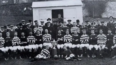Peebles Rovers A large group of football players, along with coaches, line up in a black-and-white photograph from 1908. A large trophy takes centre stage