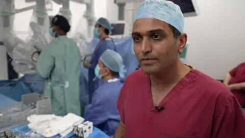Gordon Anderson/BBC A surgeon wearing a red V-neck top and a scrub cap in the foreground. There is a team of medical staff behind him surrounding a robot performing surgery.