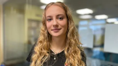 Isabelle has long wavy blonde hair and blue eyes and is wearing a black t-shirt. She is standing in front of a glass wall reflecting a row of ceiling lights.