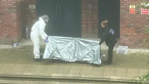 Supplied A police officer and a forensics officer holding a small silver tent in front of a brick building.