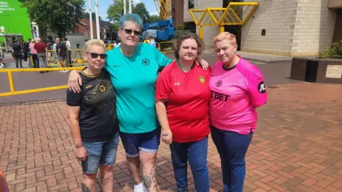 Four women in different coloured football shirts - black, blue, red, and pink - stand in front of the Molineux football ground together.