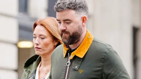 PA Media Lauren King, who has long red hair and is wearing a green coat, walks arm-in-arm with Ben King, who has dark hair peppered with grey and a brown beard. They both look sombre. 