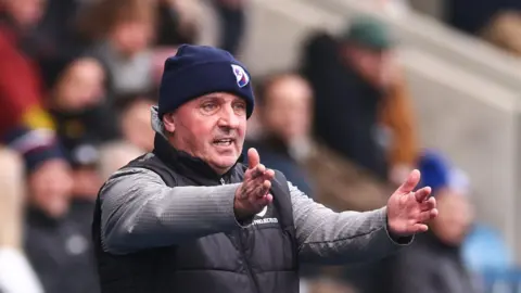 Paul Cook, wearing a woolly hat and black gilet, has his arms outstretched as he shouts instructions to his players