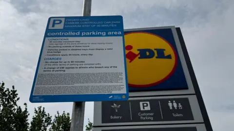 A blue parking notice beside a Lidl sign sets out the car park's terms and conditions. The sign states there is a 90-minute maximum stay, that no parking is permitted outside store hours and that a £90 charge may apply for breaching the conditions. The signs are pictured against a cloudy sky with trees in the background.