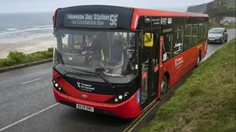 PA Media A red single-decker bus going along a costal road with a beach behind. The bus is the number 6 going to Newquay Bus Station via Constantine Bay,