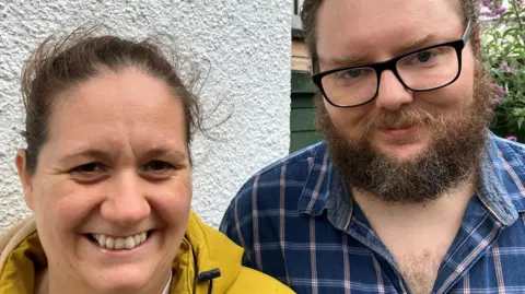 Molly and Gary Casling outside the community larder. Milly has pulled-back brown hair and is wearing a yellow coat. Gary has brown hair and a beard, and is wearing glasses and a blue checked shirt   