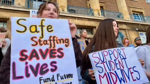 Nikki Fox/BBC Two student midwives stand on the steps of city hall in Norwich holding banners. One is colourful and reads "safe staffing saves lives" the other in blue and red simply says "support student midwives". One of the students has long brown hair which is worn down, the other has mousy blonde hair tied back in a pony tail.