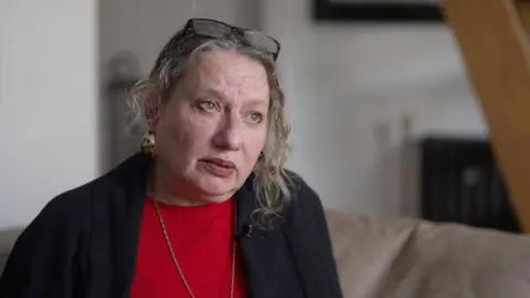 Deborah, a pistillate   with just  curly hairsbreadth  and her glasses perched connected  her head, wearing a navy jacket, a reddish  jumper and a bladed  golden  chain, sitting connected  a sofa