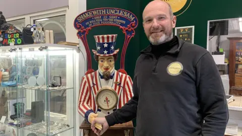 BBC/Lucy Ashton A man with a bald head and short beard in a black jumper is smiling. He is holding the hand of a statue of Uncle Sam with a stars and stripe hat and jacket. The statue is a fortune telling machine which asks people to shake its hand.