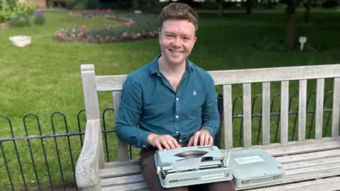 A man in brown trousers and navy shirt sits on a wooden bench, an old typewriter on his knee. Behind him are low iron railings, grass, and a flower bed.