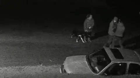 Wiltshire Police Black and white CCTV footage of a man helping another man stand up while a woman with a dog watches on. There is a a car in the road.