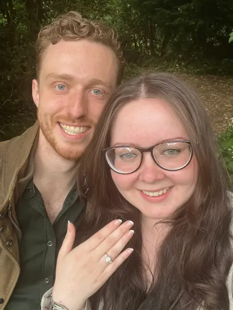 Cordelia Sheridan A couple stand in a wooded area. The woman on the left has brown hair and glasses. She is holding her hand in the air showing an engagement ring on her finger. The man on the left has brown curly hair and blue eyes. Both of them are smiling into the camera.