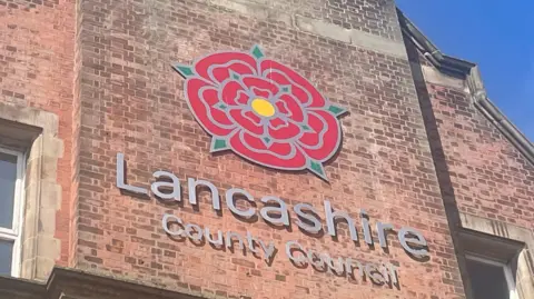 A sign of a large red rose symbolising the county of Lancashire with Lancashire County Council written beneath in white lettering. It's set against a backdrop of a red brick wall, with windows to the left and right. The sky in the picture is blue.