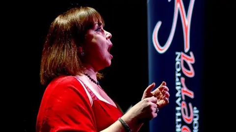 John Lloyd Davies A solo vocalist in a red dress performs on a dark stage, positioned in profile and gesturing with raised hands. A tall banner beside the performer displays the text “Northampton Opera Group”.
