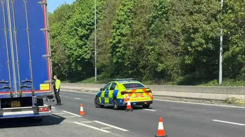 Nick Foster Blue HGV parked with police vehicle and a male police officer nearby. Two traffic cones are on the road. There are trees in the background.