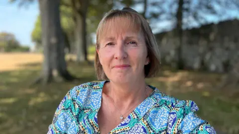 BBC Close-up outdoor portrait of a woman, who is Liz Ortan, with short, light-brown hair, centered in the frame and looking toward the camera. The woman is wearing a blue, green, and white patterned top and a silver necklace with a small pendant. The background is softly blurred, showing a row of trees, a grassy area, and a stone wall in natural daylight. The image has a shallow depth of field, keeping the person in sharp focus while the surroundings are out of focus.