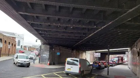 The underneath of a bridge with large steel beams above it. There are cars queueing in a line underneath it, as well as a van waiting on the left