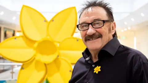 Marie Curie Barry Chuckle smiling looking straight on. He is wearing a black shirt with a yellow daffodil flower badge. He is stood next to a large daffodil-shaped statue. 