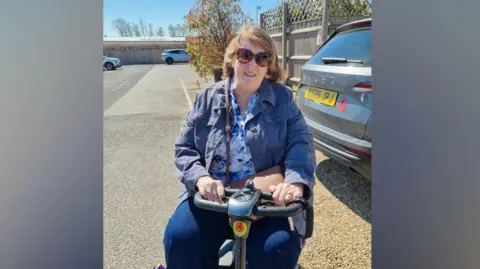 Stella Cleary A woman with light brown hair sitting on a mobility scooter in a car park. She is wearing sunglasses, a navy jacket and a blue floral patterned top with dark blue trousers. 