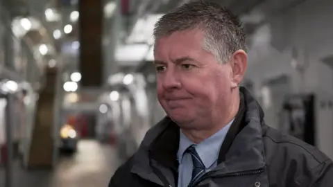 BBC Head and shoulders image of Jim Beatson, deputy governor at HMP Barlinnie. He has short, grey hair and is wearing a grey waterproof jacket with light blue shirt and dark blue tie