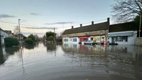 Steve Berman A flooded residential road along a parade of shops, with the water flowing all the way down the road 