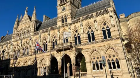 Kate Bradbrook/BBC The Guildhall, a large ornate Victorian brick building, with arched windows, arches and two flags flying outside. The building is over two floors. 