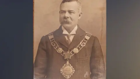 Thetford Town Council Allan Minns in about 1904, in a sepia-toned black and white photo. He is has short greying hair and a large bristling moustache, covering his upper lip. He is wearing a jacket and underneath a wing-tipped white shirt. Over his jacket is a mayoral chain. It has two rows of links and a large medallion dangling across his stomach