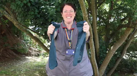 Royal United Hospitals Bath A woman in a grey short-sleeved NHS uniform with rainbow lanyard is smiling broadly to camera and holding a pair of long blue socks in front of her. She is standing in front of a tree with several low branches